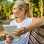Blonde Frau mit Tablet in der Hand auf Parkbank sitzend